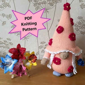 May include: A pink knitted gnome with a white lace trim hat and pink flowers. The gnome is holding a blue flower in one hand. The image includes the text "PDF Knitting Pattern".
