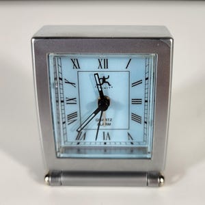 May include: A silver-colored quartz alarm clock with a light blue face and black hands. The clock face features Roman numerals and the word "QUARTZ ALARM". The clock has a rectangular shape with a curved base.