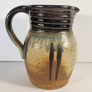 May include: Handmade ceramic pitcher with a dark brown handle and rim. The body of the pitcher features a speckled, earthy color palette with streaks of brown and blue. The pitcher has a classic, rustic design.