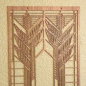 May include: A wooden wall art piece with a geometric Art Deco design. The artwork features symmetrical patterns of lines and shapes, creating a stylized, abstract representation of a tree or plant. The piece is made of light brown wood and has a rectangular shape.