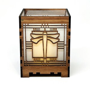May include: A wooden candle holder with a dragonfly design. The square-shaped holder is made of light brown wood and features a laser-cut dragonfly in the center. The dragonfly is surrounded by geometric patterns. The interior has a soft, glowing light.