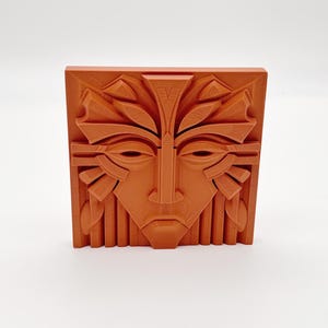 May include: A terracotta-colored, square relief sculpture featuring a stylized face with geometric and organic elements. The face has almond-shaped eyes, a prominent nose, and a detailed headdress. The artwork is likely a decorative piece.