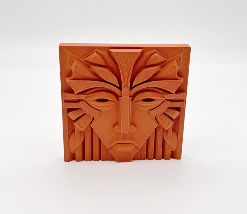 May include: A terracotta-colored, square relief sculpture featuring a stylized face with geometric and organic elements. The face has almond-shaped eyes, a prominent nose, and a detailed headdress. The artwork is likely a decorative piece.