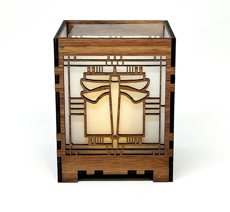 May include: A wooden candle holder with a dragonfly design. The square-shaped holder is made of light brown wood and features a laser-cut dragonfly in the center. The dragonfly is surrounded by geometric patterns. The interior has a soft, glowing light.
