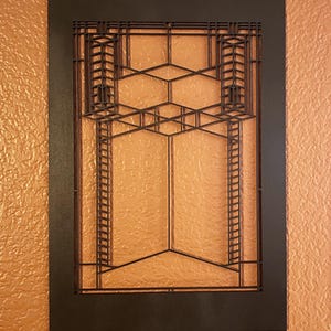 May include: A dark brown, geometric wall art piece with a rectangular frame. The design features a series of intersecting lines and shapes, creating a layered, architectural pattern. The artwork is mounted on a textured, orange-toned wall.