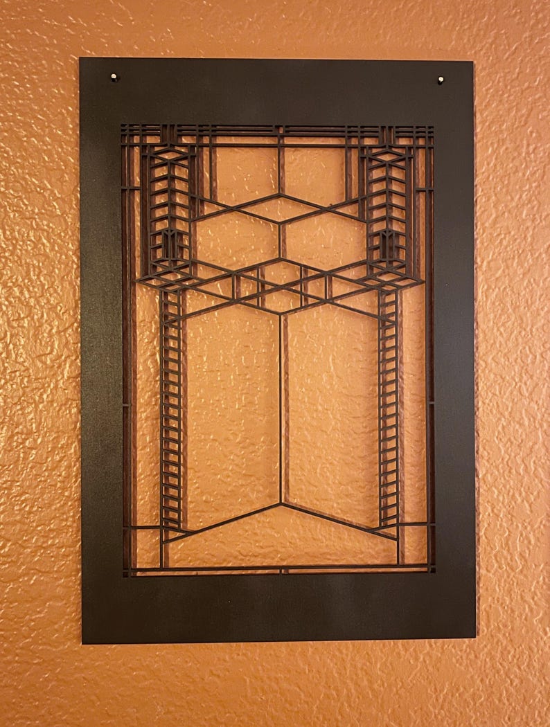 May include: A dark brown, geometric wall art piece with a rectangular frame. The design features a series of intersecting lines and shapes, creating a layered, architectural pattern. The artwork is mounted on a textured, orange-toned wall.