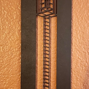 May include: A black metal wall art piece with a geometric design. The art features a rectangular frame with a central cutout showcasing a ladder-like structure against a textured orange background. Decorative home decor.