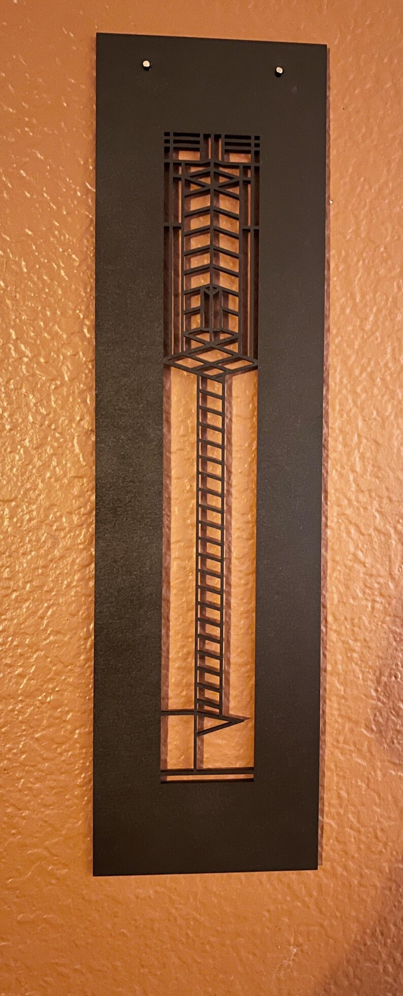 May include: A black metal wall art piece with a geometric design. The art features a rectangular frame with a central cutout showcasing a ladder-like structure against a textured orange background. Decorative home decor.