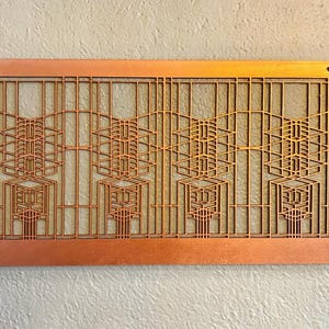 Puede incluir: Una pieza de arte mural rectangular de color cobre con un dise&ntilde;o geom&eacute;trico. La obra de arte presenta un patr&oacute;n repetitivo de formas rectangulares y angulares, creando un efecto de capas. Se ven dos peque&ntilde;os agujeros en la parte superior para colgar. La pieza est&aacute; montada en una pared clara.