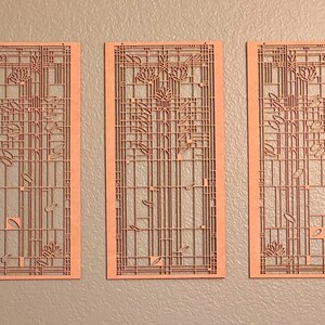 May include: Three laser-cut wooden panels with a floral and geometric design. The panels are rectangular with a light brown colour and feature intricate cut-out patterns of flowers, leaves, and vertical and horizontal lines. The panels are displayed against a grey wall.