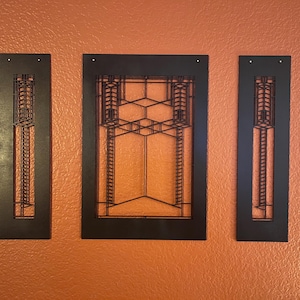 May include: Three-piece black metal wall art set. The set includes two vertical rectangular pieces and one larger rectangular piece with a geometric design. The art is mounted on an orange textured wall.