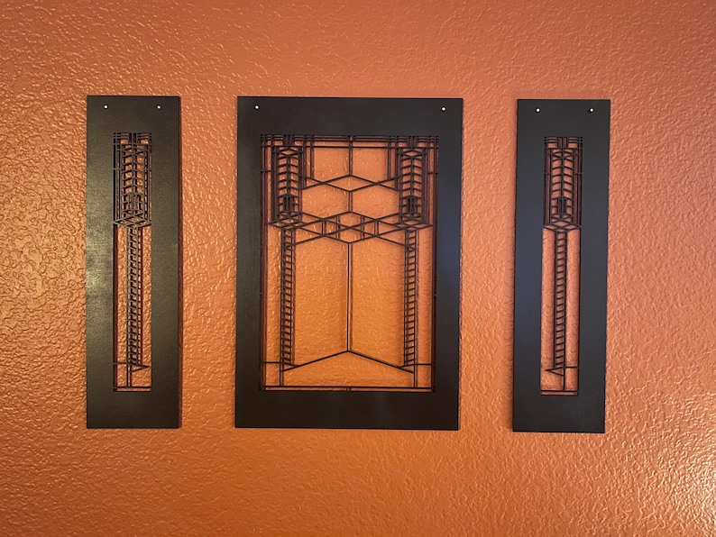 May include: Three-piece black metal wall art set. The set includes two vertical rectangular pieces and one larger rectangular piece with a geometric design. The art is mounted on an orange textured wall.