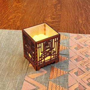 May include: A square, wooden candle holder with an intricate geometric design. The candle holder is illuminated from within, casting a warm glow. It sits on a patterned fabric runner with a wooden surface in the background.