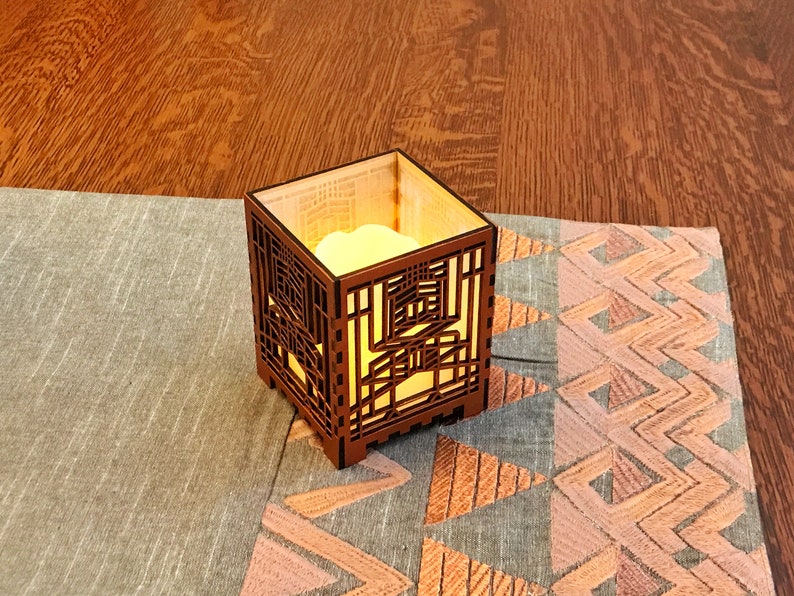 May include: A square, wooden candle holder with an intricate geometric design. The candle holder is illuminated from within, casting a warm glow. It sits on a patterned fabric runner with a wooden surface in the background.