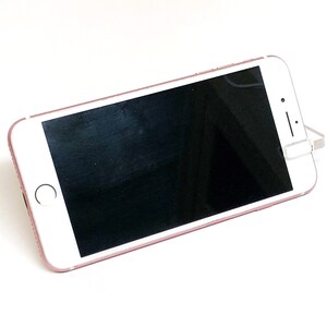 May include: A rose gold smartphone with a black screen is propped up on a white surface. The phone has a white front with a circular home button and a camera. The phone is angled slightly to the right.
