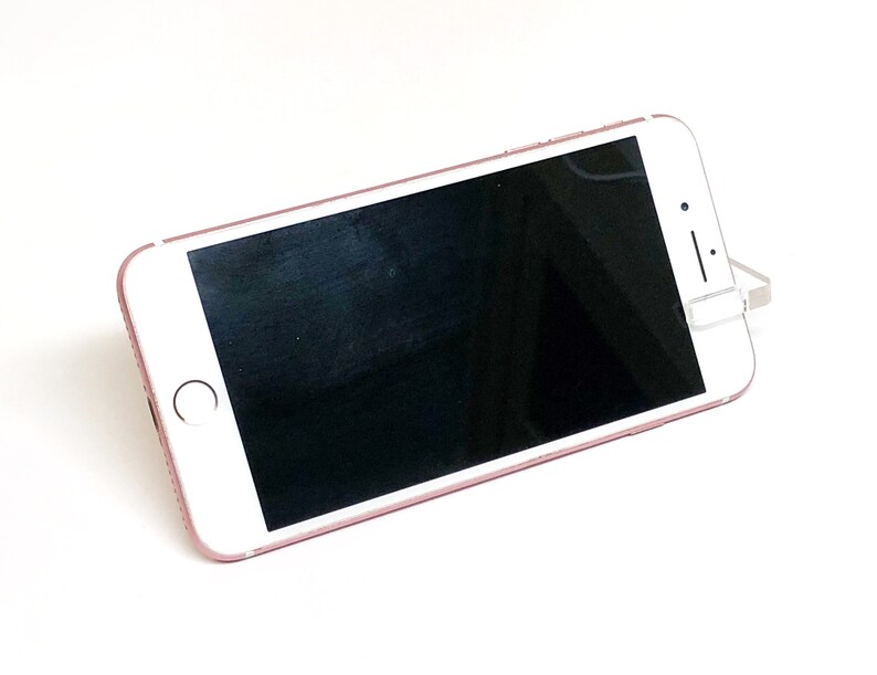 May include: A rose gold smartphone with a black screen is propped up on a white surface. The phone has a white front with a circular home button and a camera. The phone is angled slightly to the right.