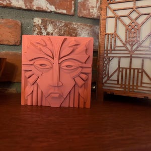 May include: A coral-colored, square relief sculpture featuring a stylized face with leaf-like details. Next to it is a wooden, geometric-patterned lamp with a warm, glowing interior. The objects are displayed on a dark wooden surface.
