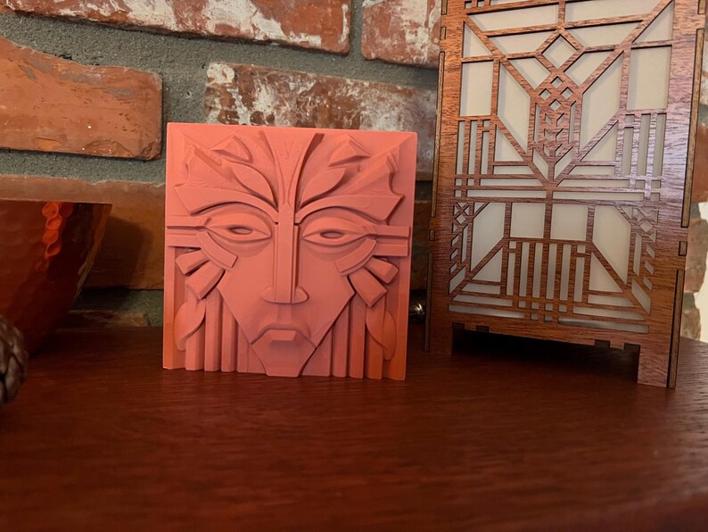 May include: A coral-colored, square relief sculpture featuring a stylized face with leaf-like details. Next to it is a wooden, geometric-patterned lamp with a warm, glowing interior. The objects are displayed on a dark wooden surface.