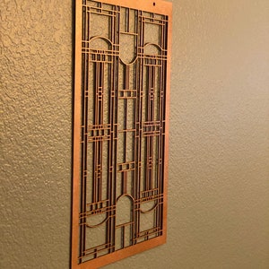 Op de afbeelding: Een koperkleurig, rechthoekig kunstwerk voor aan de muur met een geometrisch ontwerp. Het kunstwerk heeft een reeks verticale en horizontale lijnen, waardoor een rasterachtig patroon ontstaat met gebogen en rechthoekige vormen. Twee kleine gaten zitten aan de bovenkant om op te hangen.