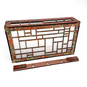 May include: A rectangular, decorative wooden wall panel with a geometric design. The panel features a series of rectangular and square cutouts, with a copper-coloured frame and a distressed, aged finish. A separate wooden piece with three keyhole cutouts is included.