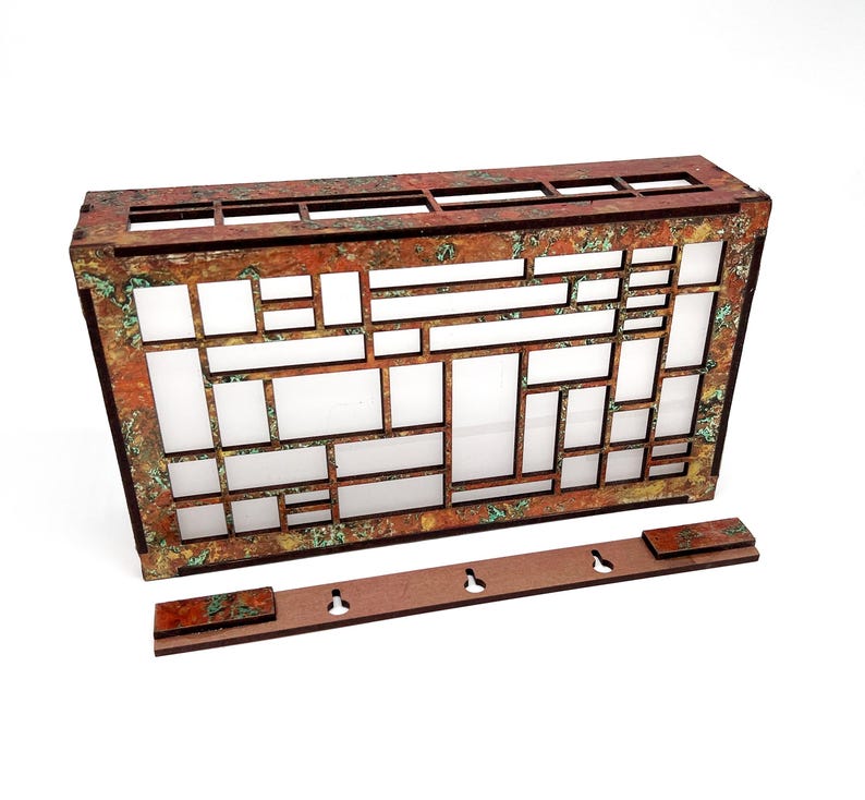 May include: A rectangular, decorative wooden wall panel with a geometric design. The panel features a series of rectangular and square cutouts, with a copper-coloured frame and a distressed, aged finish. A separate wooden piece with three keyhole cutouts is included.