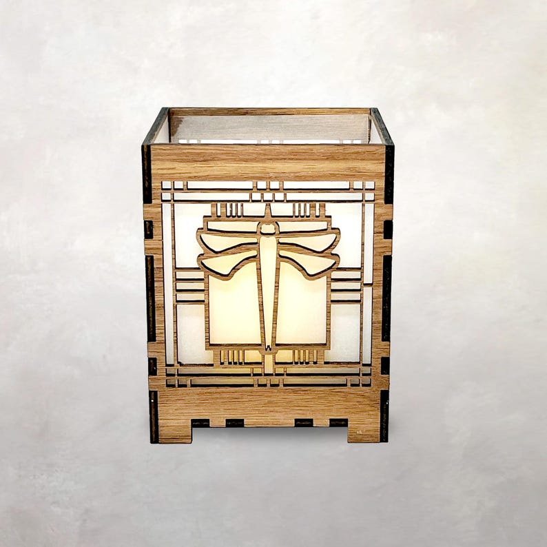 May include: A wooden lantern featuring a dragonfly design. The lantern is crafted from light-coloured wood, showcasing a geometric pattern and a dragonfly silhouette. The interior emits a soft light, creating a warm atmosphere.