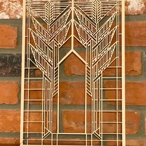 May include: A light-colored, laser-cut wooden wall art piece with an Art Deco design. The artwork features symmetrical, stylized tree-like forms and geometric patterns. The piece is rectangular with two small holes at the top for hanging.