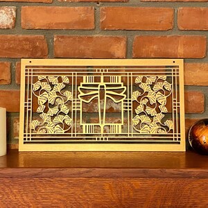 May include: A gold-colored, rectangular wall art piece with a dragonfly and ginkgo leaf design. The artwork is set against a brick wall and rests on a wooden surface. A candle and decorative sphere are also present.