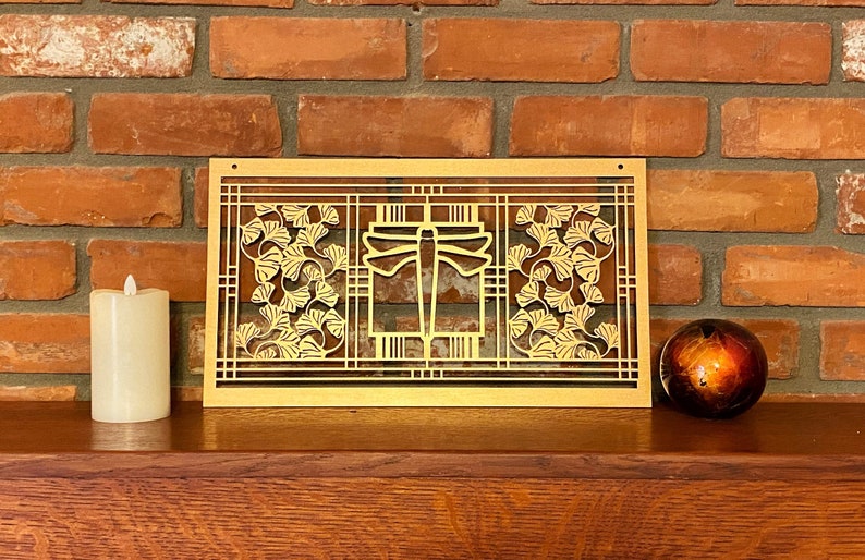 May include: A gold-colored, rectangular wall art piece with a dragonfly and ginkgo leaf design. The artwork is set against a brick wall and rests on a wooden surface. A candle and decorative sphere are also present.