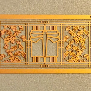 May include: A rectangular, golden-colored wall art piece with a cut-out design. The artwork features a central geometric shape flanked by panels with stylized ginkgo leaves. The piece is mounted on a textured, light gray wall.