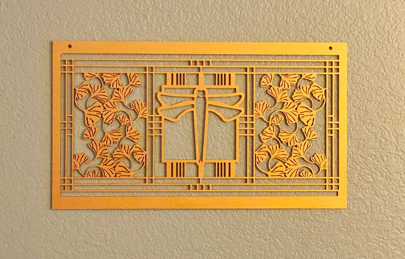 May include: A rectangular, golden-colored wall art piece with a cut-out design. The artwork features a central geometric shape flanked by panels with stylized ginkgo leaves. The piece is mounted on a textured, light gray wall.