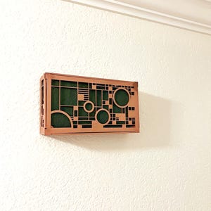 Op de afbeelding: Een geometrisch houten wandkunstwerk met een koperkleurig frame en een groen interieur. Het ontwerp heeft uitgesneden cirkels, vierkanten en lijnen. De kunst is gemonteerd op een witte muur met textuur.