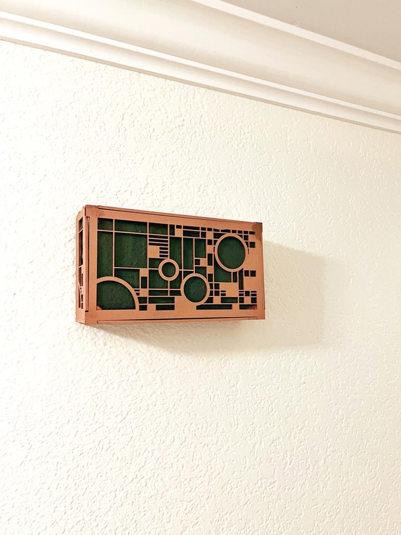 Op de afbeelding: Een geometrisch houten wandkunstwerk met een koperkleurig frame en een groen interieur. Het ontwerp heeft uitgesneden cirkels, vierkanten en lijnen. De kunst is gemonteerd op een witte muur met textuur.
