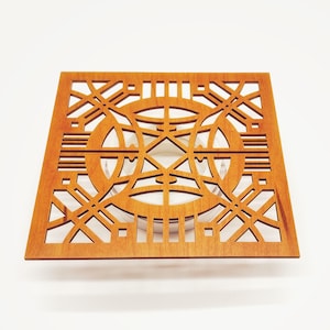 May include: A square wooden coaster with a geometric openwork design. The coaster is a warm, medium brown colour and features intersecting lines and circular shapes. The design is symmetrical and intricate, creating a decorative and functional item.