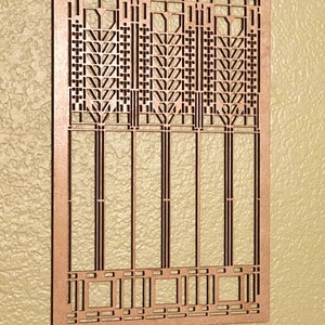 May include: A copper-colored, rectangular decorative panel with an intricate geometric design. The panel features a series of vertical and horizontal lines, creating a repeating pattern of squares, rectangles, and stylized floral motifs. Two small holes are at the top for hanging.