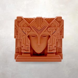 May include: A terracotta-colored, geometric art deco style sculpture of a serene face. The face is centered within a square, with elaborate architectural details framing it. The sculpture has a symmetrical design and a matte finish.