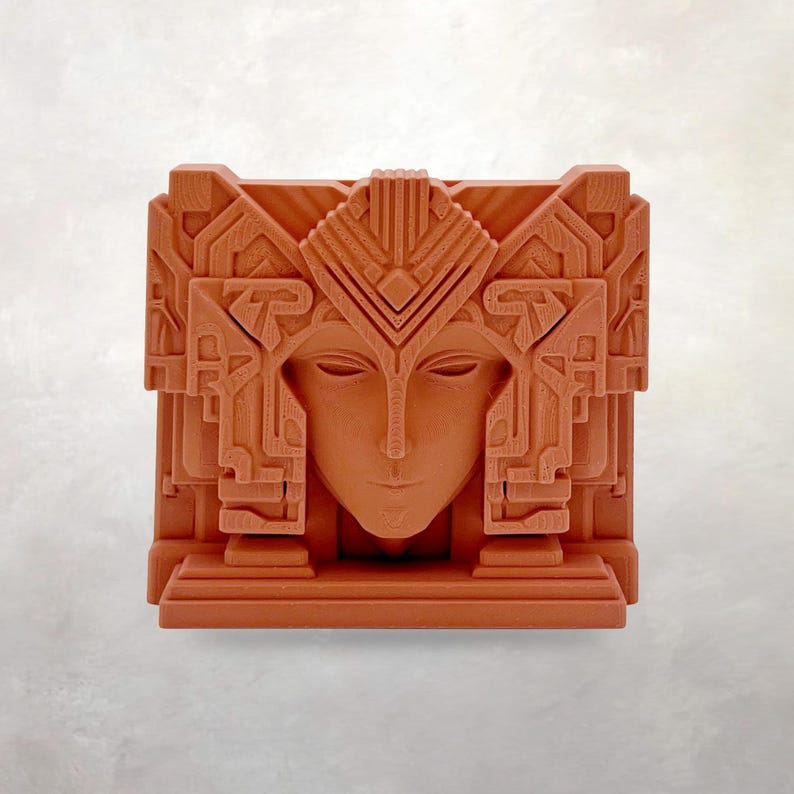 May include: A terracotta-colored, geometric art deco style sculpture of a serene face. The face is centered within a square, with elaborate architectural details framing it. The sculpture has a symmetrical design and a matte finish.