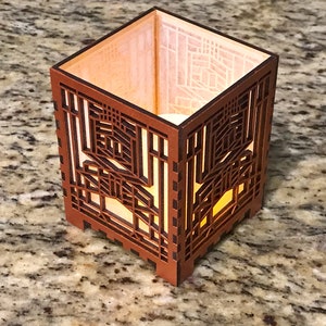 May include: A square, wooden candle holder with intricate geometric cutouts. The warm, amber glow of a light source inside illuminates the detailed design. The exterior is a rich, dark brown.