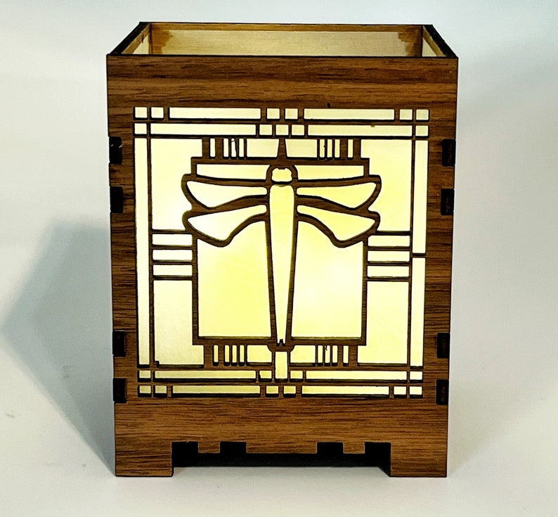 May include: A wooden box with a dragonfly design. The box is made of dark brown wood and features a dragonfly silhouette in the center, with a yellow light shining through the design. The box has a rectangular shape.