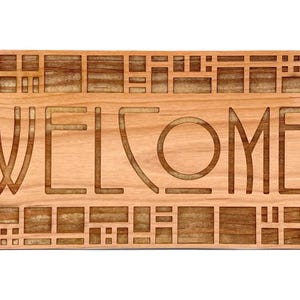 Puede incluir: Un letrero rectangular de madera con la palabra &laquo;WELCOME&raquo; tallada en la superficie. El letrero es de madera de color marr&oacute;n claro y presenta un patr&oacute;n geom&eacute;trico alrededor de los bordes y entre las letras.