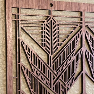 Puede incluir: Primer plano de una pieza de arte mural de madera cortada con l&aacute;ser. El dise&ntilde;o presenta un patr&oacute;n geom&eacute;trico de estilo Art D&eacute;co con intrincados recortes. La madera tiene un tono marr&oacute;n c&aacute;lido y un peque&ntilde;o agujero en la parte superior para colgar.