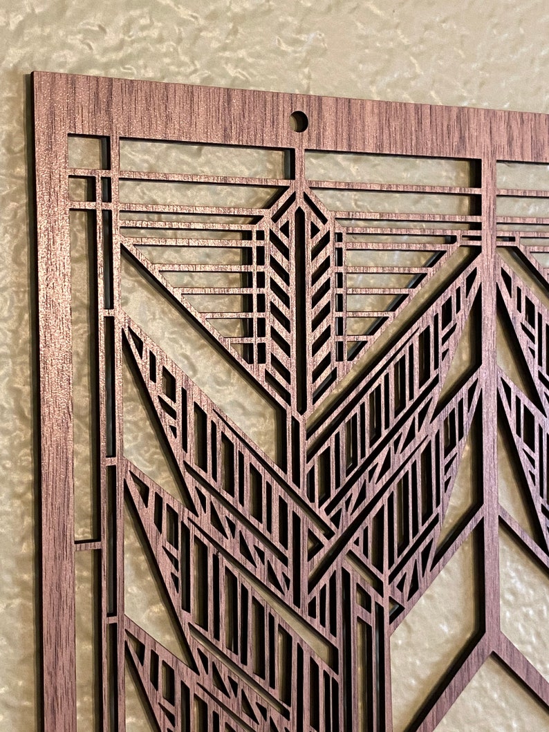 Puede incluir: Primer plano de una pieza de arte mural de madera cortada con l&aacute;ser. El dise&ntilde;o presenta un patr&oacute;n geom&eacute;trico de estilo Art D&eacute;co con intrincados recortes. La madera tiene un tono marr&oacute;n c&aacute;lido y un peque&ntilde;o agujero en la parte superior para colgar.