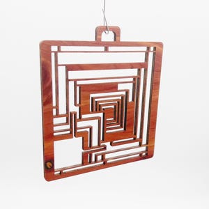 May include: A square, wooden ornament with a geometric design. The ornament is made of reddish-brown wood and features a series of rectangular and L-shaped cutouts, creating a layered, abstract pattern. A small loop is at the top for hanging.