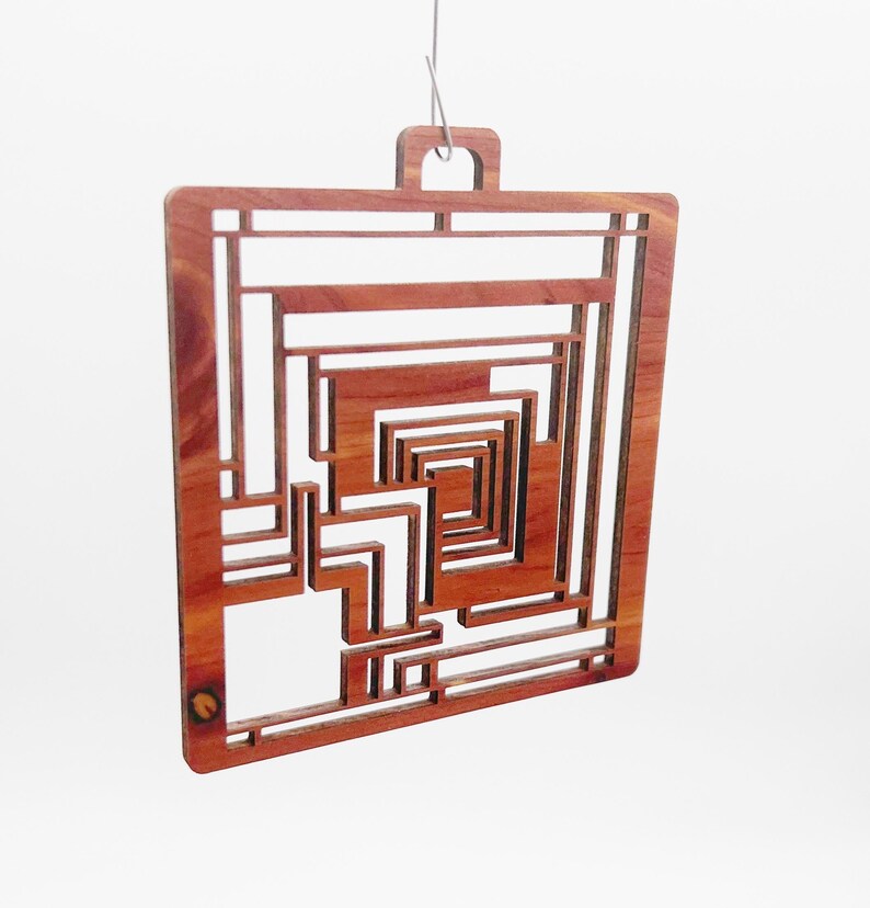 May include: A square, wooden ornament with a geometric design. The ornament is made of reddish-brown wood and features a series of rectangular and L-shaped cutouts, creating a layered, abstract pattern. A small loop is at the top for hanging.