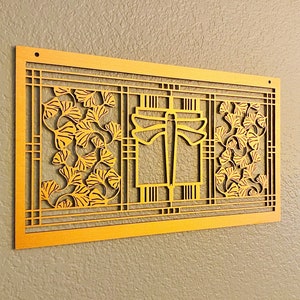 May include: A gold-colored, rectangular wall art piece with a dragonfly and ginkgo leaf design. The artwork features a layered, cut-out design with intricate details. The piece is mounted on a neutral-colored wall, showcasing its decorative nature.