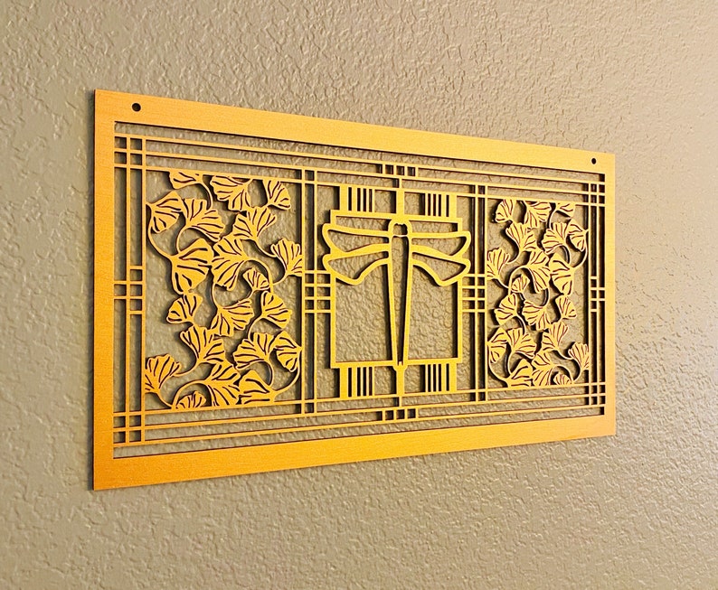 May include: A gold-colored, rectangular wall art piece with a dragonfly and ginkgo leaf design. The artwork features a layered, cut-out design with intricate details. The piece is mounted on a neutral-colored wall, showcasing its decorative nature.