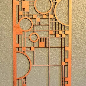 Op de afbeelding: Een koperkleurig metalen wandkunstwerk met een geometrisch ontwerp met vierkanten, rechthoeken en cirkels. Het ontwerp doet denken aan de Art Deco-stijl.