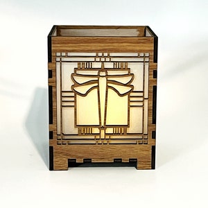 May include: A wooden box featuring a dragonfly design. The box is constructed from light brown wood and has a rectangular form. The dragonfly is centrally positioned, set against a white backdrop. The box is illuminated internally, producing a warm light.