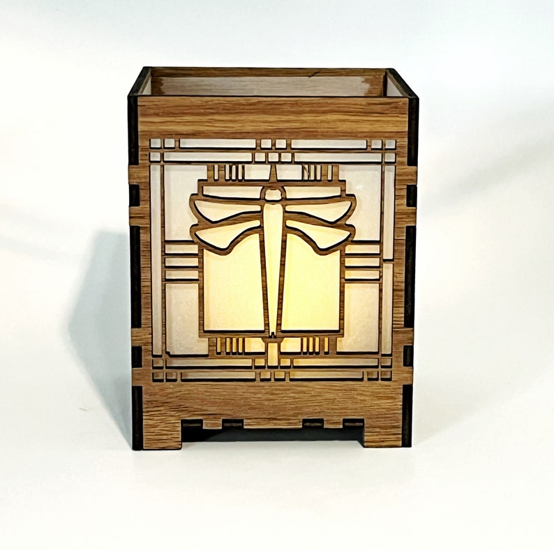May include: A wooden box featuring a dragonfly design. The box is constructed from light brown wood and has a rectangular form. The dragonfly is centrally positioned, set against a white backdrop. The box is illuminated internally, producing a warm light.
