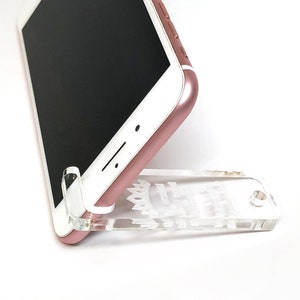 May include: A rose gold smartphone rests on a clear acrylic phone stand. The stand is designed to hold the phone upright. The phone's screen is black, and the stand has decorative cutouts.